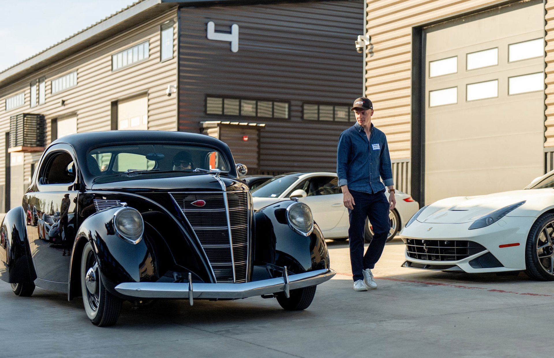 Owner walking past a classic black coupe and Ferrari outside Unit 4 at GOTA